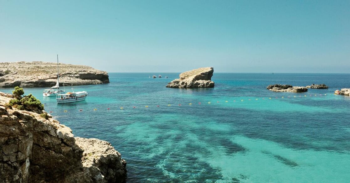 A serene coastal landscape featuring crystal-clear turquoise waters lapping against rugged, sun-drenched cliffs. Several boats are anchored in the calm bay, with a few rocky outcrops dotting the expansive blue sea under a clear sky.