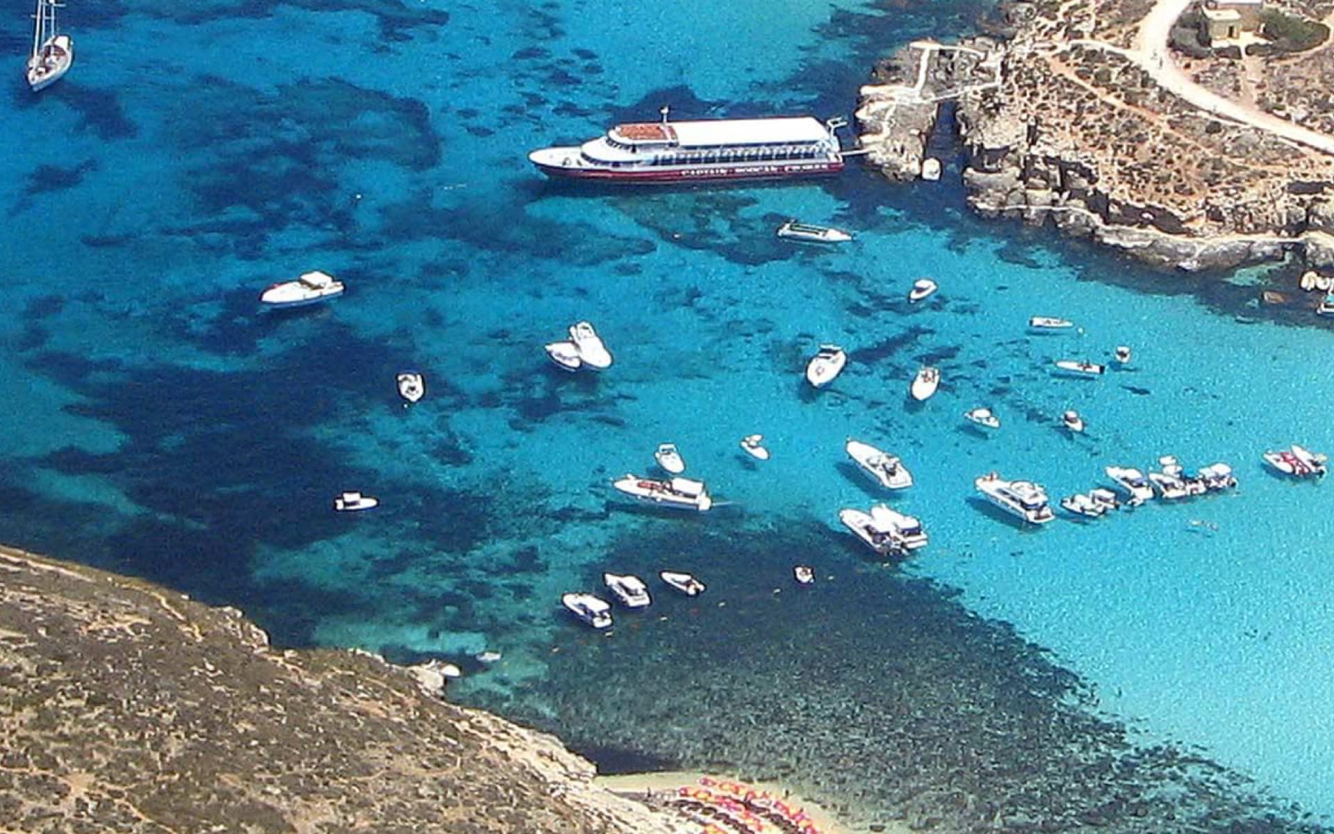 A picturesque aerial view of a bay filled with crystal-clear turquoise waters, dotted with numerous boats and yachts anchored near a rocky coastline.