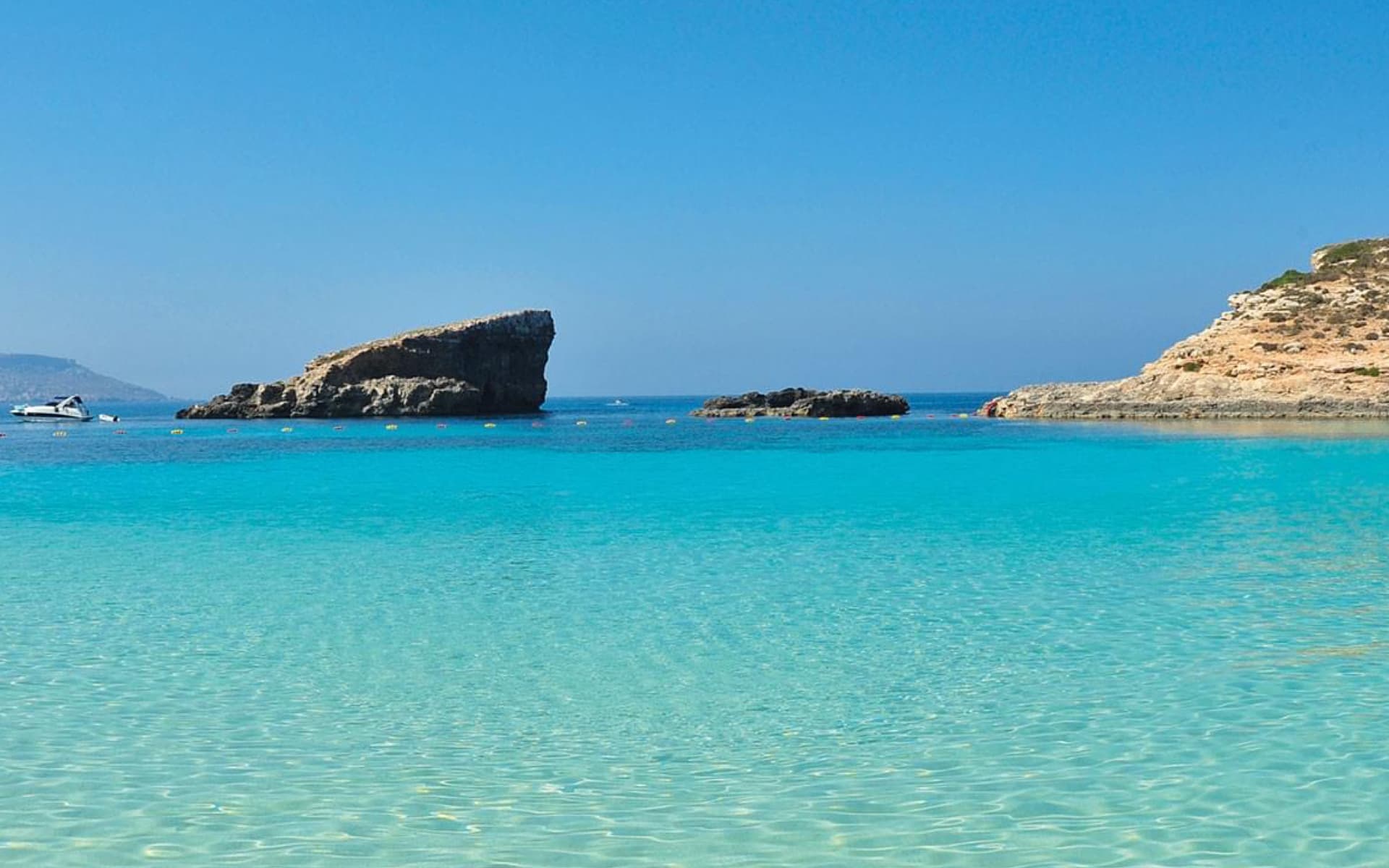 The image shows a stunning, crystal-clear turquoise water beach with rocky formations and a boat in the distance under a bright blue sky.
