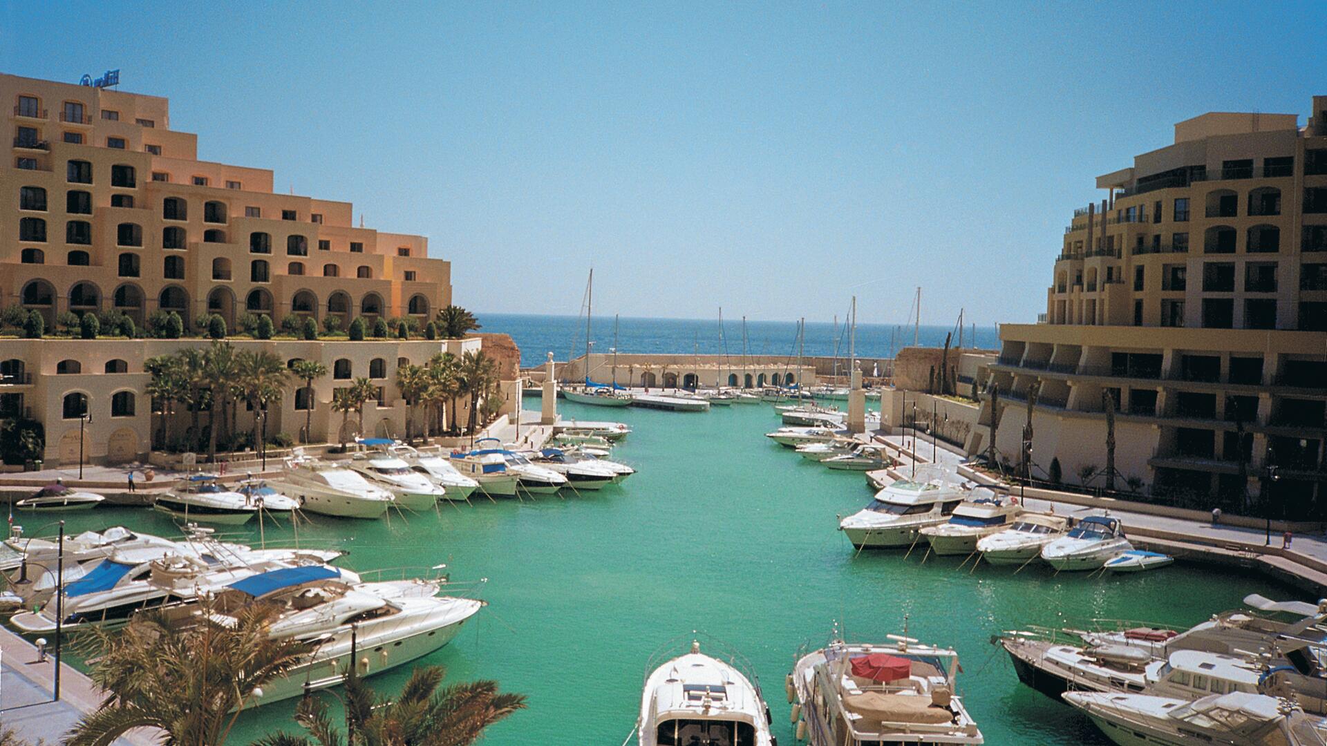 This is a scenic view of a marina filled with numerous boats and yachts, nestled between large, multi-story buildings with a distinct architectural style. The turquoise water reflects the clear blue sky, creating a picturesque and tranquil atmosphere.