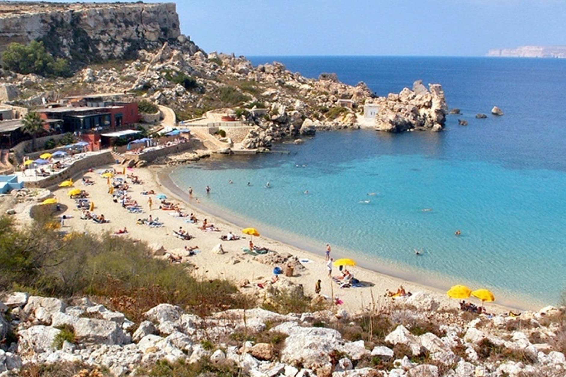 A sunny day at a busy sandy beach with turquoise waters, rocky cliffs, and numerous sunbathers relaxing under umbrellas.