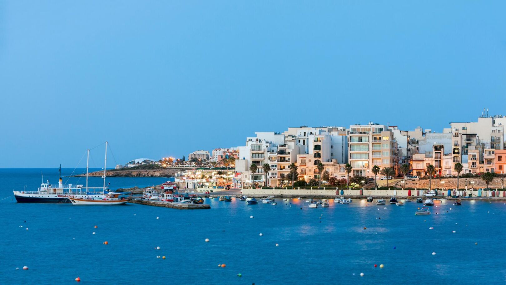 This is a scenic view of a coastal town at dusk, with buildings illuminated by lights and several boats docked in the calm blue water.