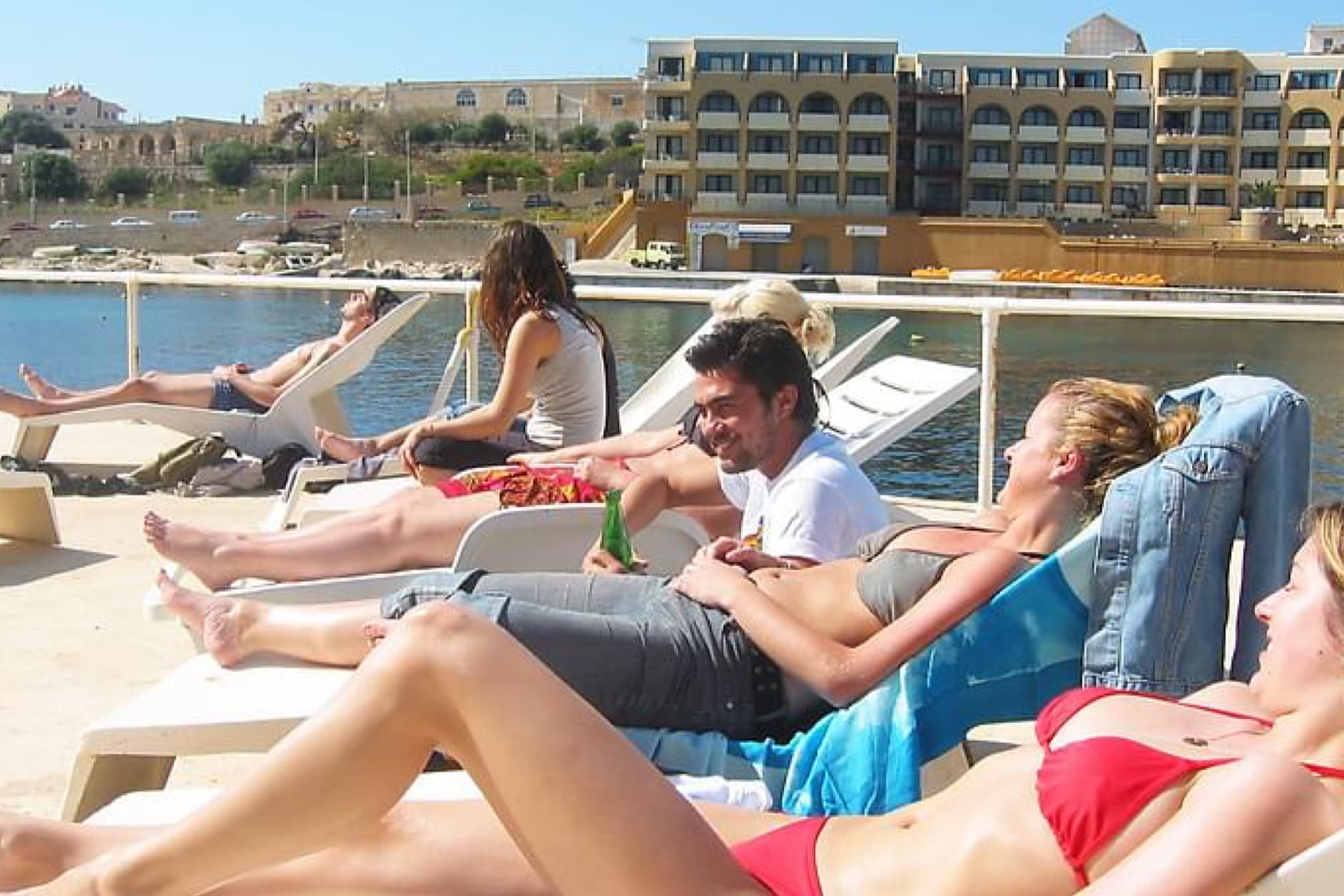 Several people are relaxing on lounge chairs on a sunny day near a body of water, with buildings in the background.