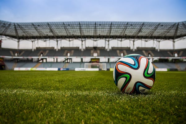 A soccer ball rests on the green grass of an empty stadium, poised for the next game. The stands are visible in the background.