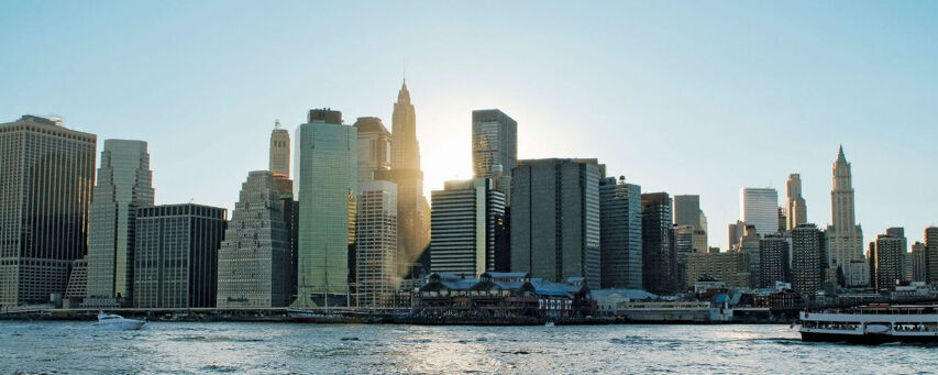 The Manhattan skyline at sunset, with the East River in the foreground and boats passing by.