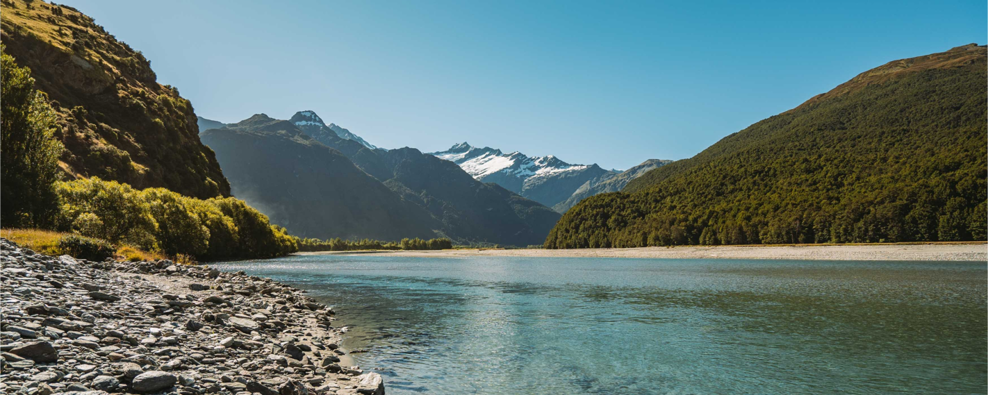 A breathtaking panoramic view of a serene lake, framed by lush forests and snow-capped mountains, with a rocky shore in the foreground.