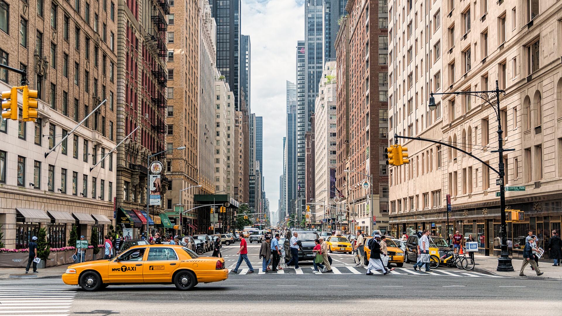 A bustling New York City street scene unfolds, with iconic yellow taxis navigating traffic amidst pedestrians crossing a busy intersection, all framed by towering skyscrapers.
