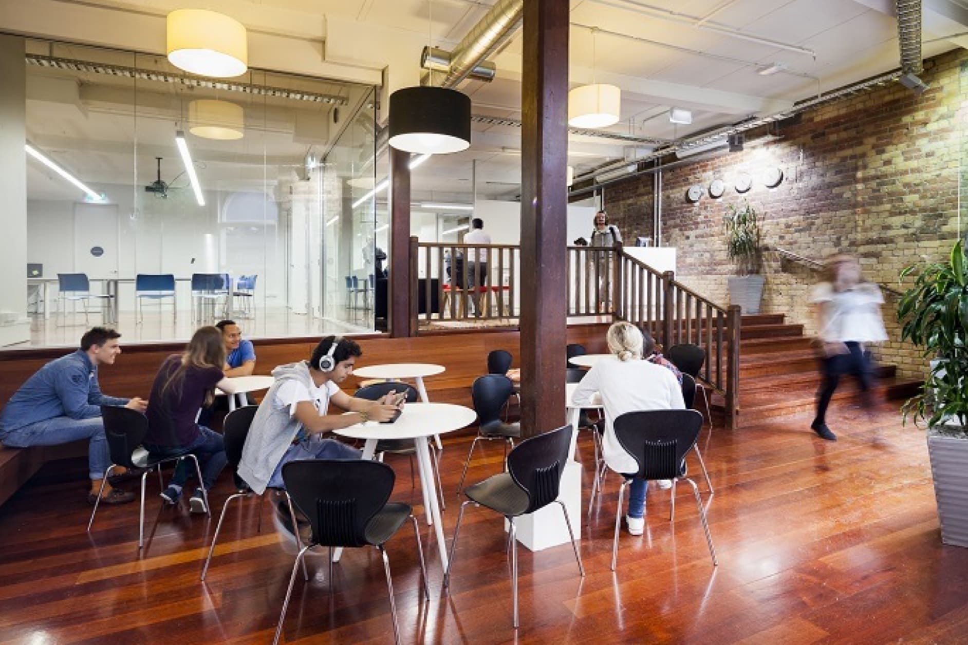 Several young adults are seated at tables in a modern, open-plan office space featuring wooden floors and brick walls.