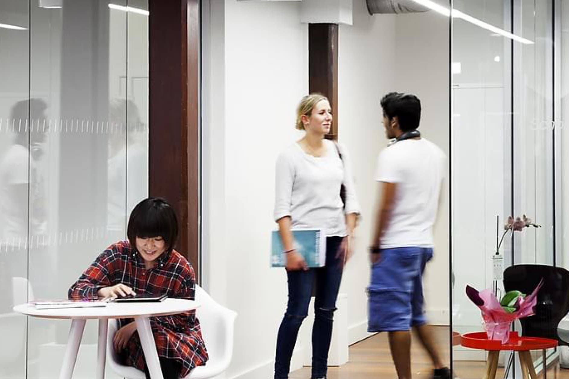 Three people are in a modern office setting. One person is working on a laptop at a small table, while two others are standing and talking near a glass partition.
