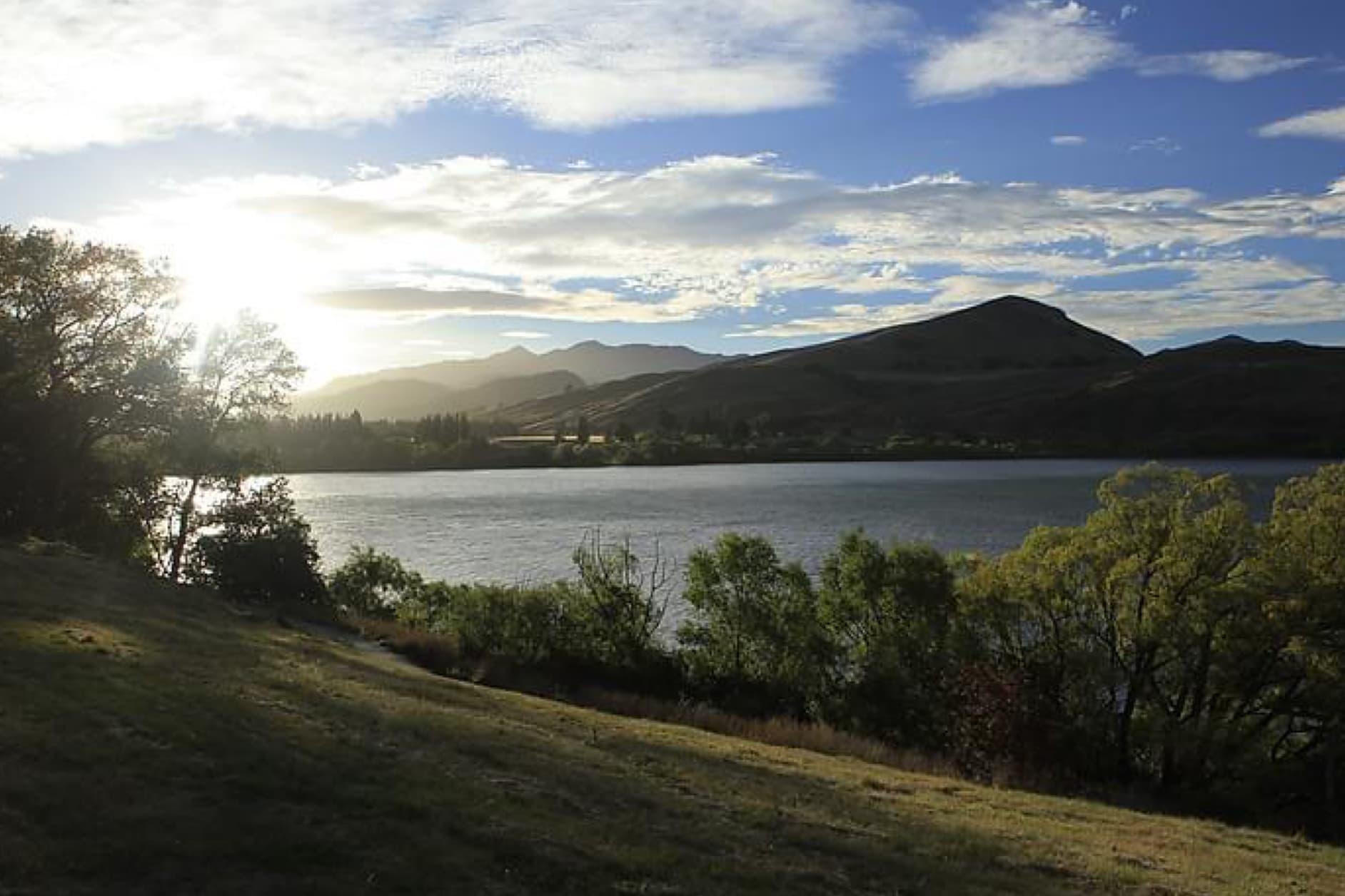 A picturesque view of a serene lake, surrounded by rolling hills and trees, with the sun shining through the clouds, casting a warm glow on the landscape.