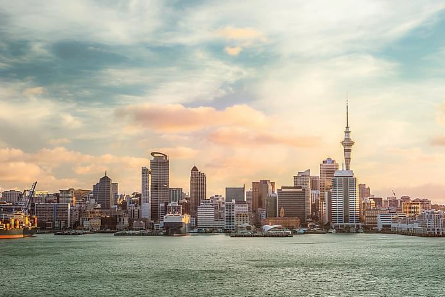 The Auckland, New Zealand skyline, featuring the Sky Tower, stretches across the water under a dramatic sky.