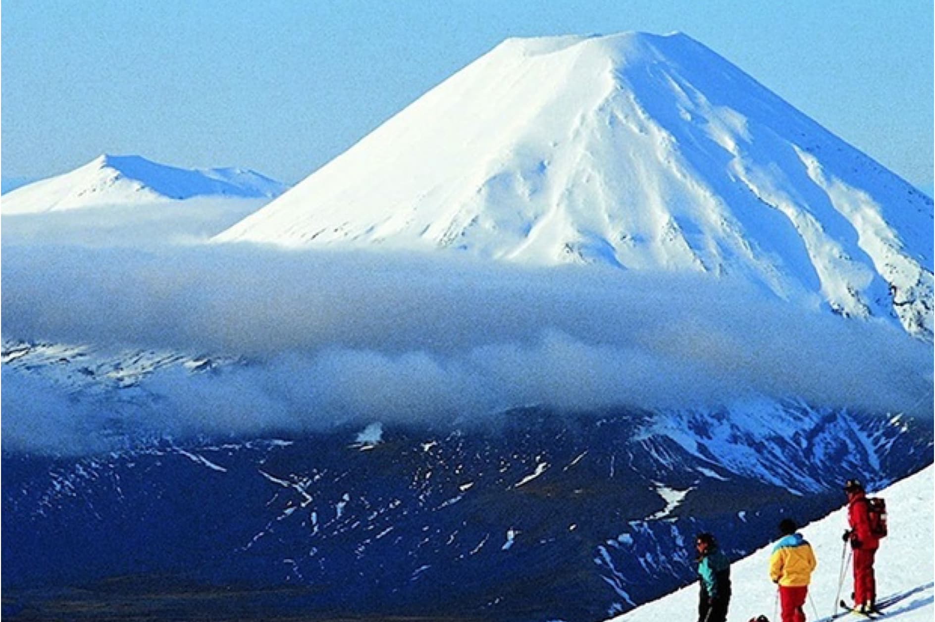 A group of skiers are enjoying a sunny day on a snow-covered mountain, with a majestic, cloud-shrouded volcano in the background.