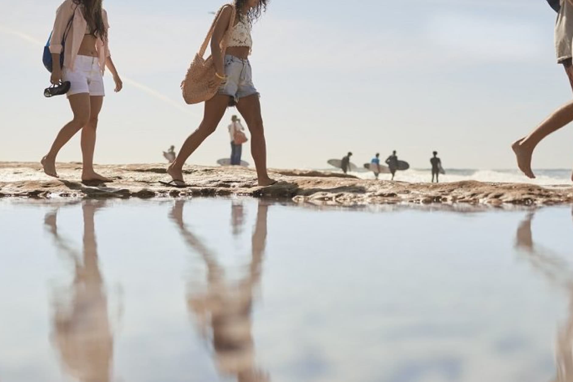 Young people enjoy a sunny day at the beach, with some strolling along the shore and others waiting for waves with their surfboards.