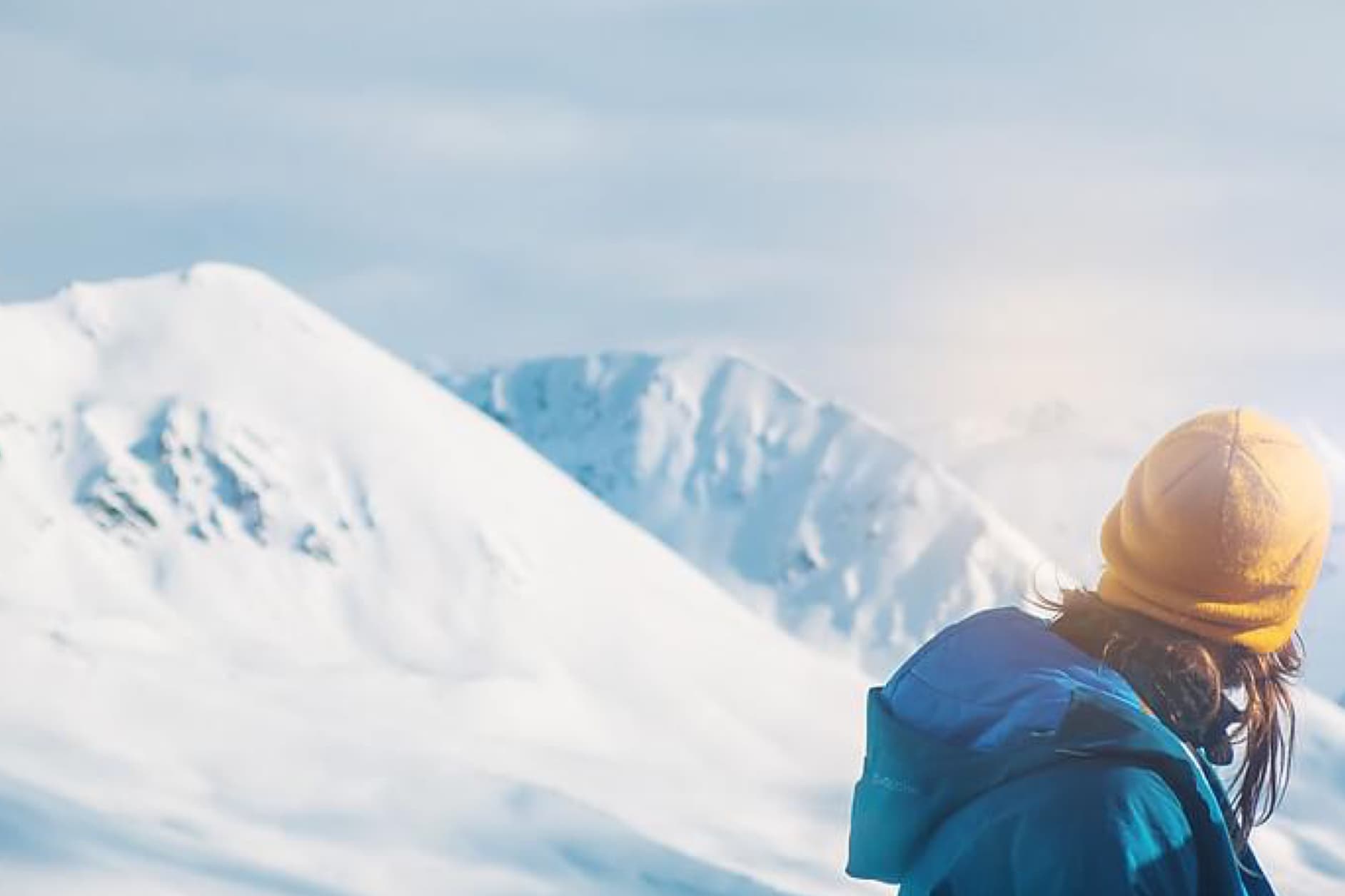 A person wearing a blue jacket and a yellow beanie looks out at a snow-covered mountain range, illuminated by the winter sun.