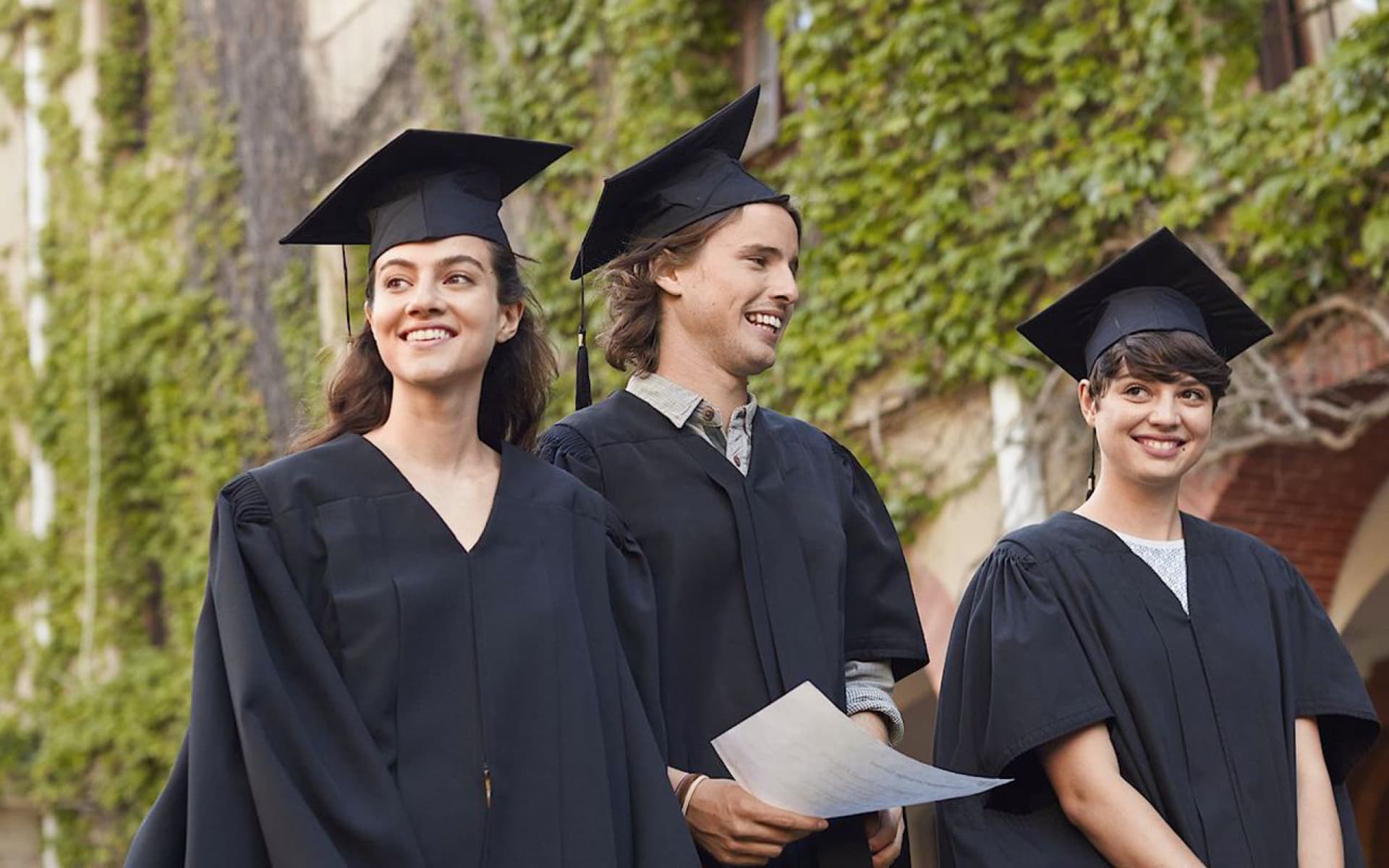 Three smiling graduates, dressed in traditional black academic gowns and caps, stand outdoors in front of a building covered in ivy. One is holding a document.