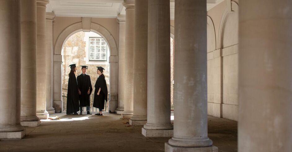 Three individuals wearing graduation caps and gowns stand in a columned hallway, engaged in conversation. The setting appears to be a historic building, likely a university.