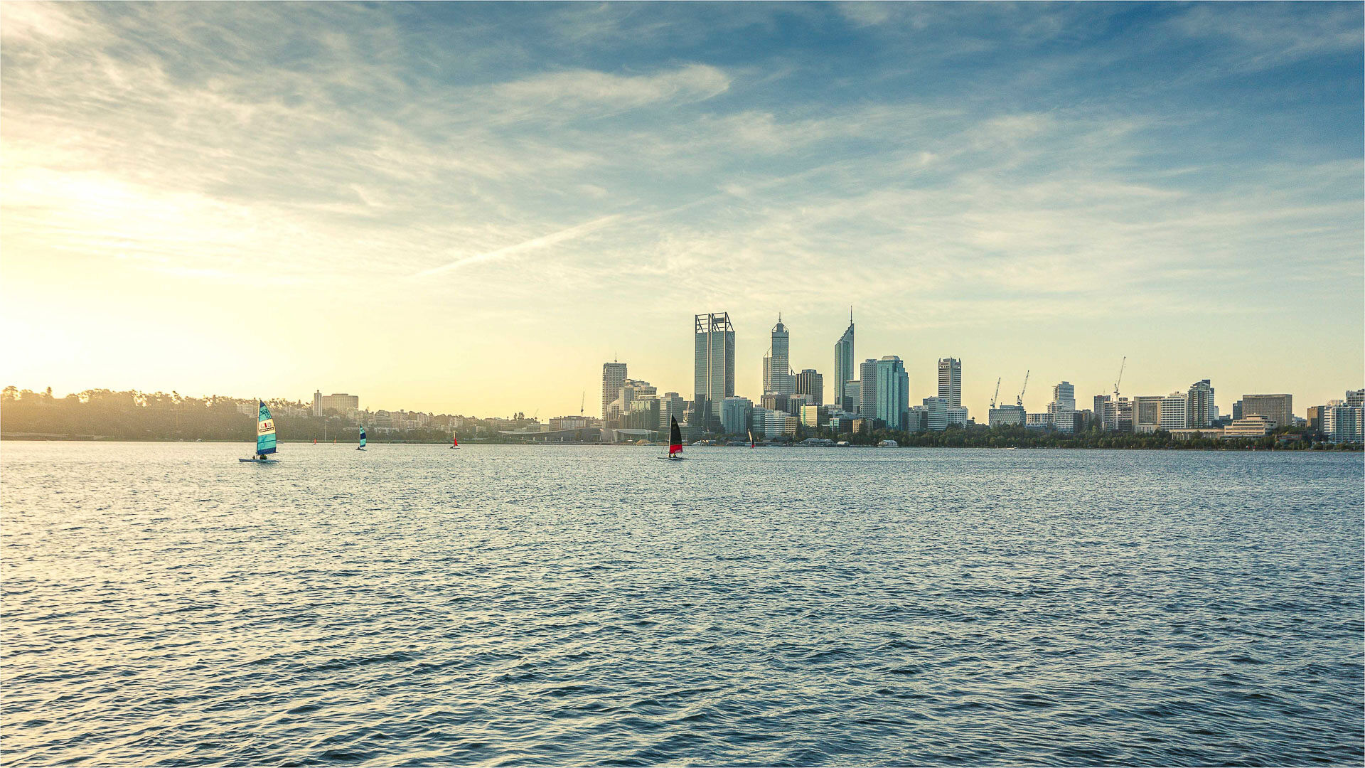 A picturesque view of the Perth, Australia skyline at sunset, with sailboats visible on the Swan River in the foreground.