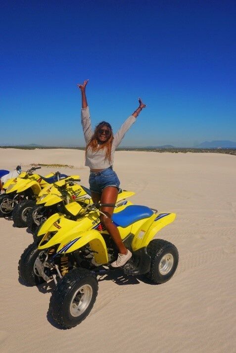 A woman is posing on a yellow ATV in a sandy desert landscape under a clear blue sky. She has her arms raised in excitement.