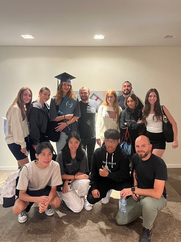 A group of 12 people, including a graduate wearing a cap and gown, pose for a photo indoors.