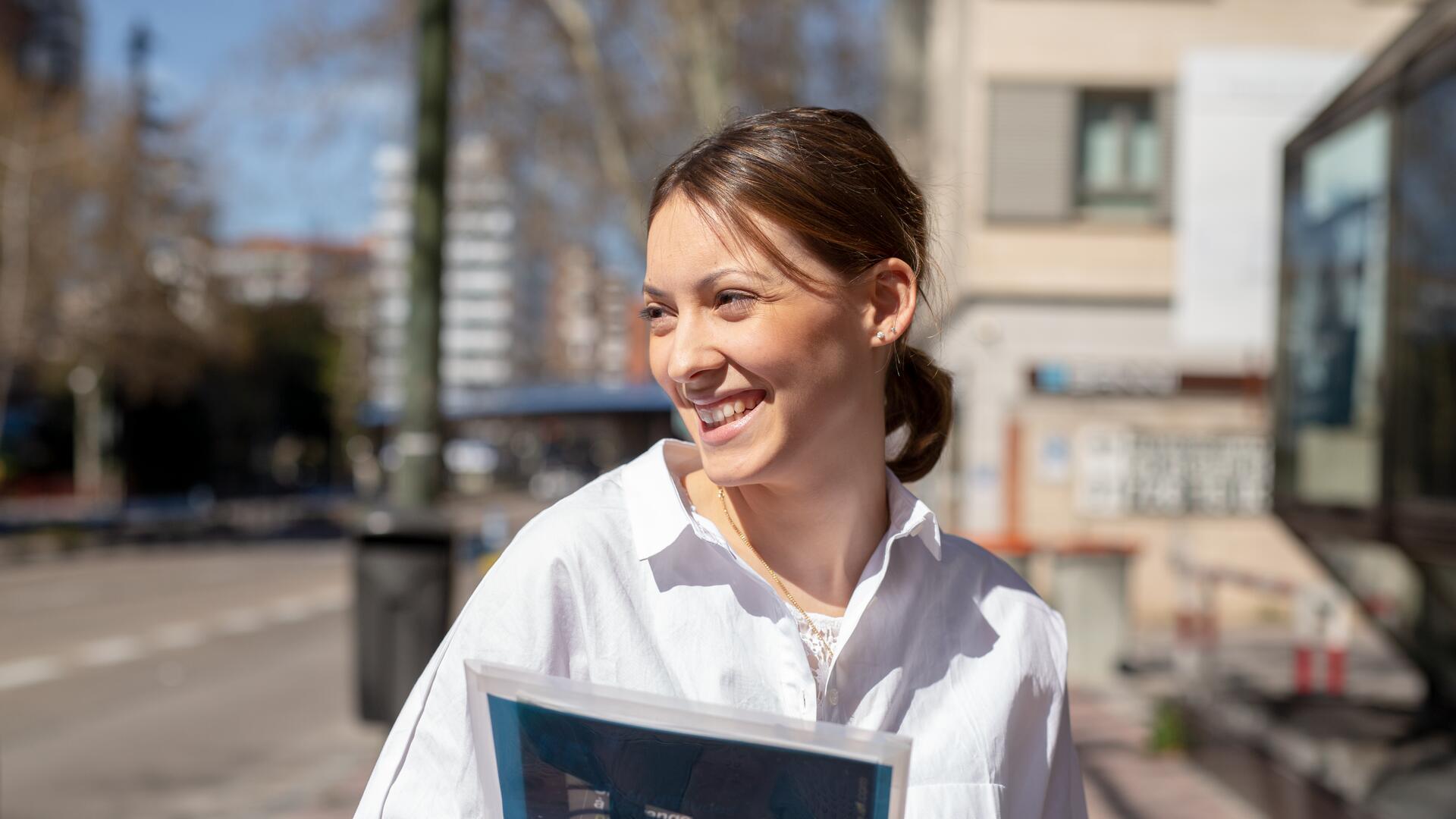 A young woman with brown hair, wearing a white coat, smiles and looks to her left while holding a folder. She is outdoors on a sidewalk with buildings in the background.