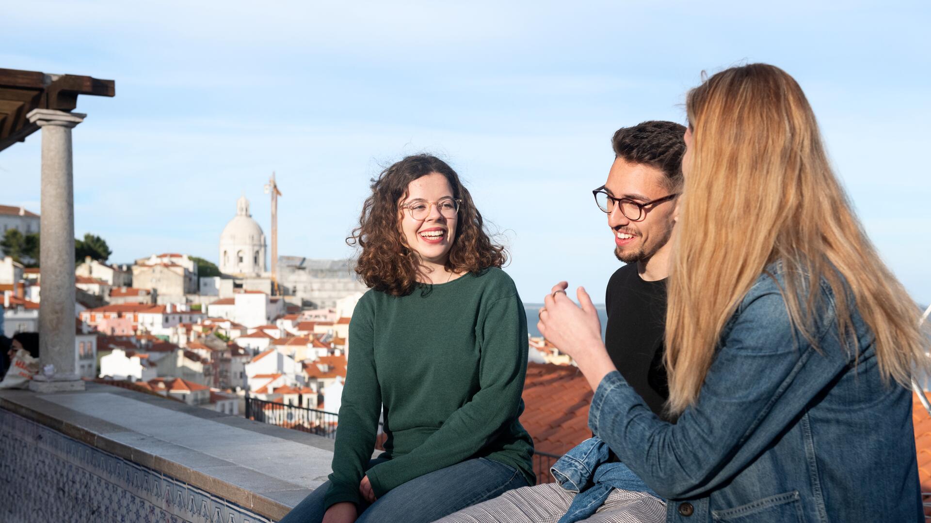 Three young adults are sitting on a ledge, enjoying a conversation with a cityscape in the background. The woman on the left has curly brown hair and glasses, wearing a green sweater. The man in the middle has dark hair and glasses, wearing a black shirt. The woman on the right has long blonde hair, wearing a denim jacket.