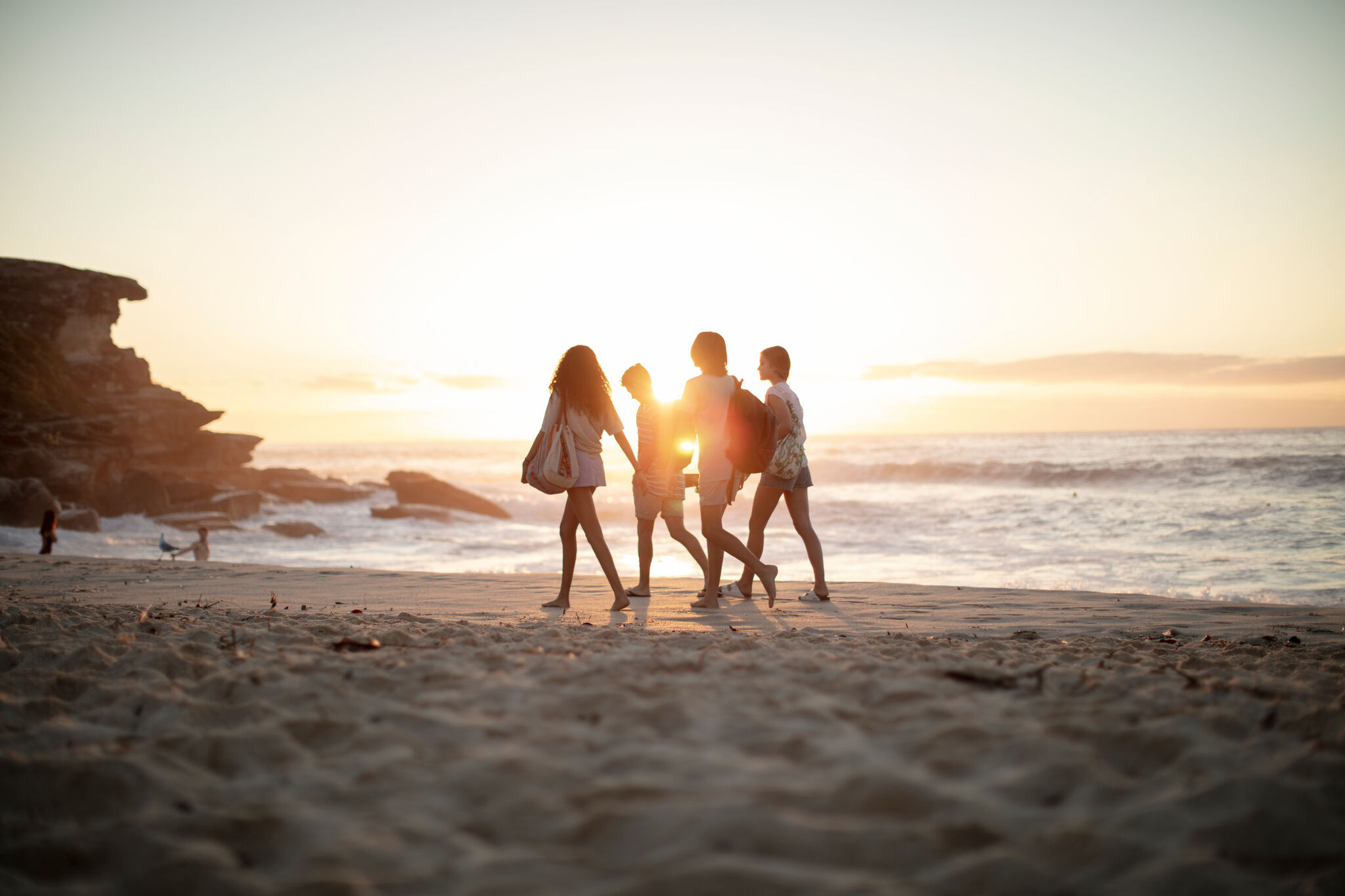 Four young women enjoy a sunny day at the beach, walking along the water's edge as the sun sets, casting a warm, golden glow.