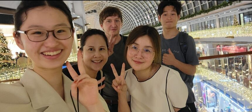 Five people pose for a selfie in a shopping mall decorated with lights. Two women in the foreground are making peace signs.