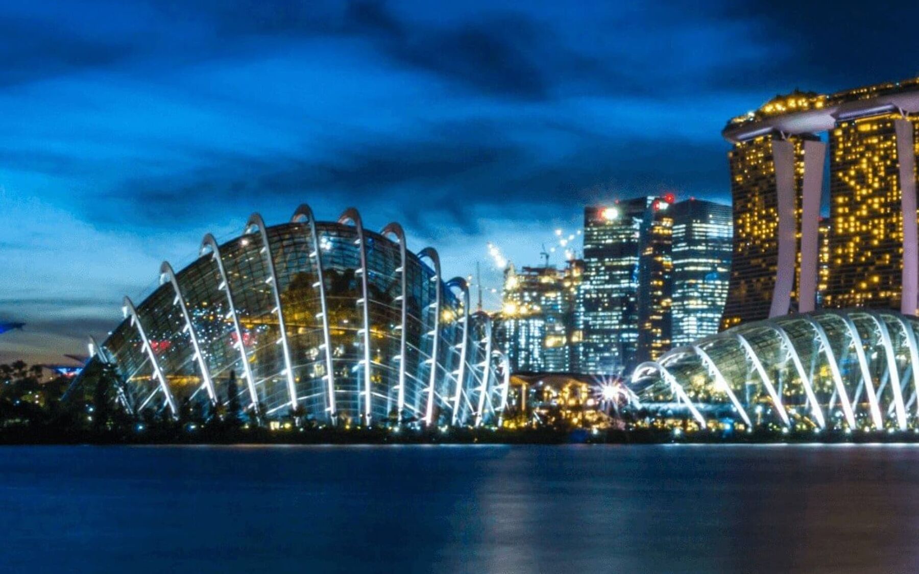 The image shows the Gardens by the Bay in Singapore at dusk, with the iconic Marina Bay Sands hotel in the background. The futuristic conservatories are illuminated, creating a stunning visual spectacle against the twilight sky.