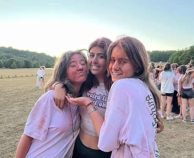 Three young women with colorful powder paint on their faces and clothes pose happily for the camera, with a group of people visible in the background.