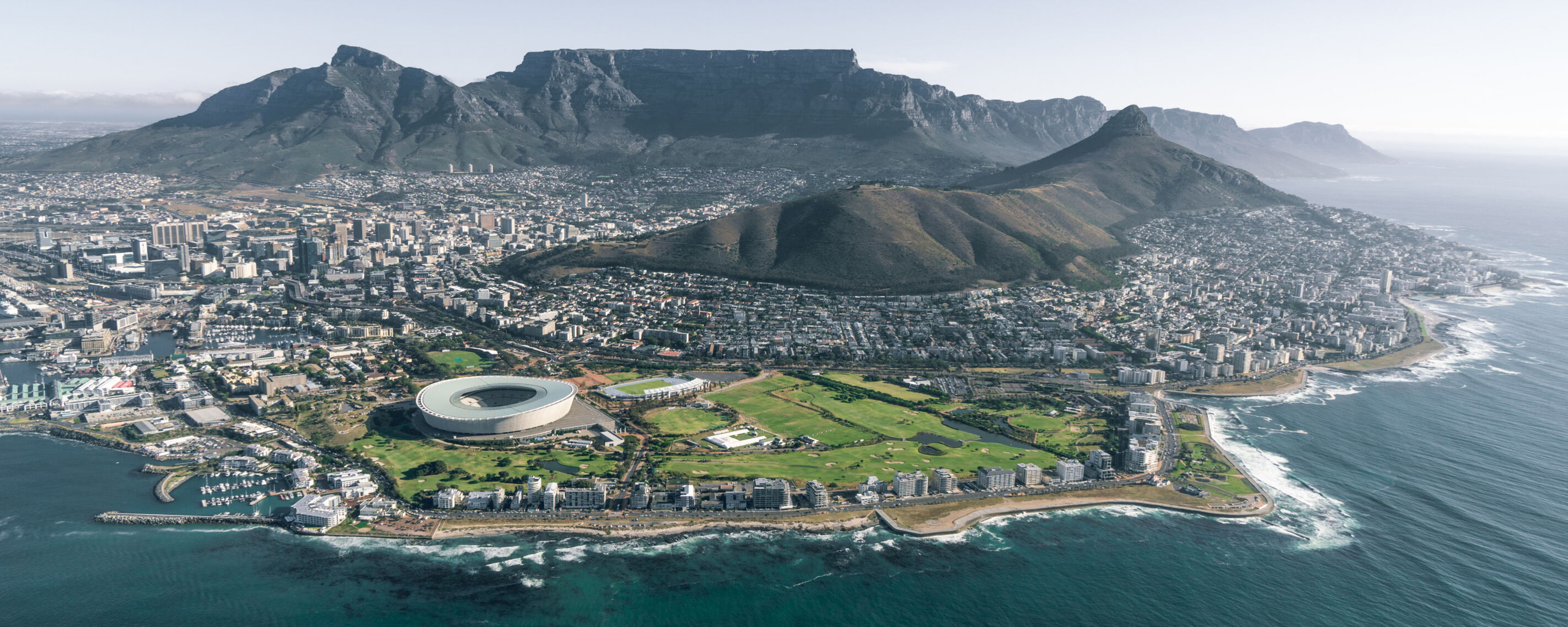 A panoramic view of Cape Town, South Africa, featuring Table Mountain in the background, Cape Town Stadium, and the ocean in the foreground.