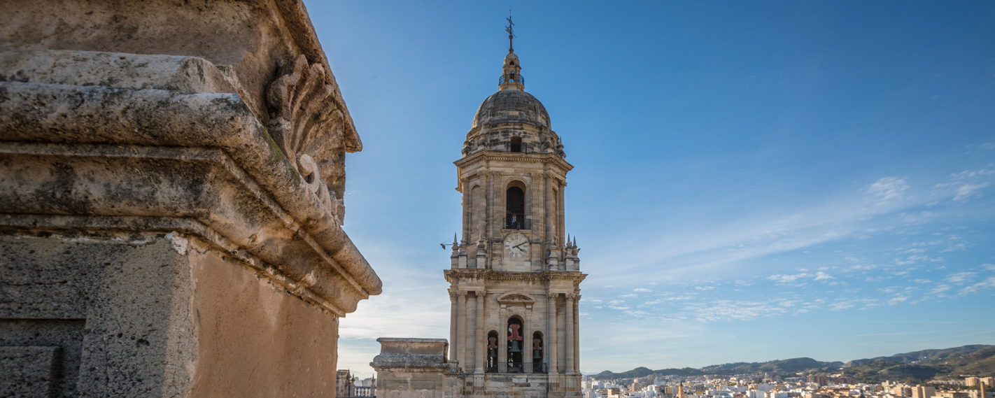 A striking view of the bell tower of Malaga Cathedral, featuring intricate stonework in the foreground and a cityscape in the background.
