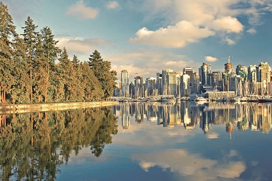 A picturesque view of the Vancouver, Canada skyline, with calm water reflecting the trees and buildings. The scene is characterized by a serene and peaceful atmosphere.