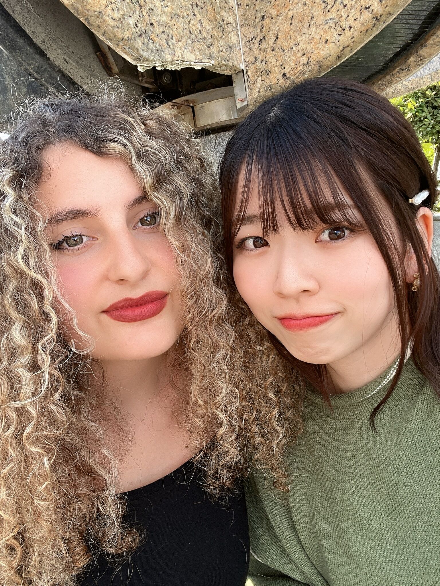 This is a close-up photo of two young women, likely friends, posing together outdoors. The woman on the left has curly blonde hair and is wearing red lipstick, while the woman on the right has dark hair and is wearing a green top.