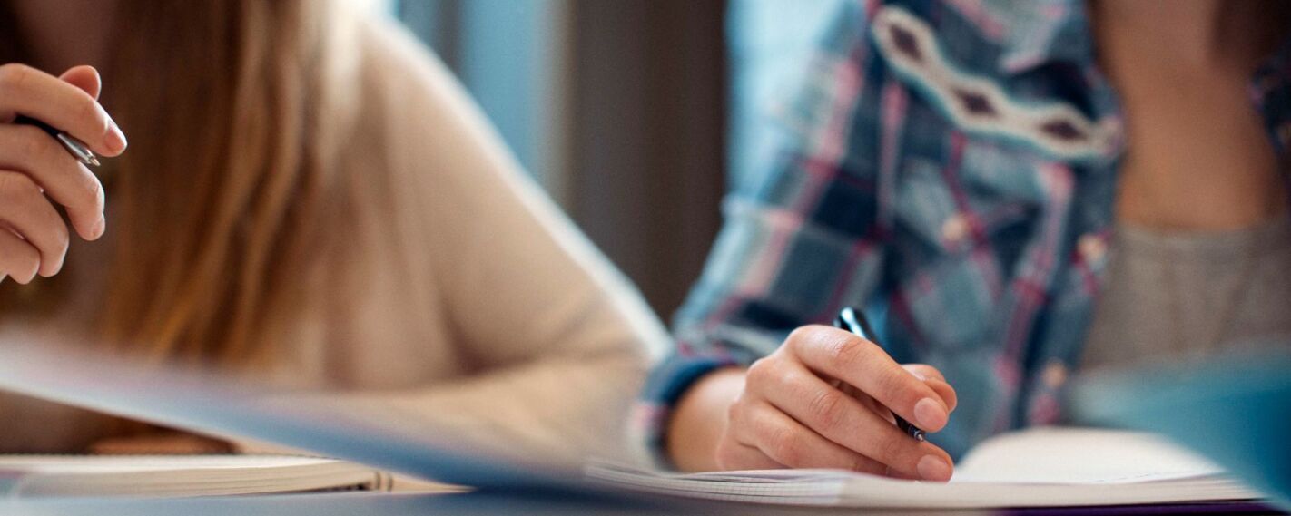 Two individuals are diligently working together, with one person holding a pen and the other actively writing in a notebook, suggesting a collaborative study or work session.