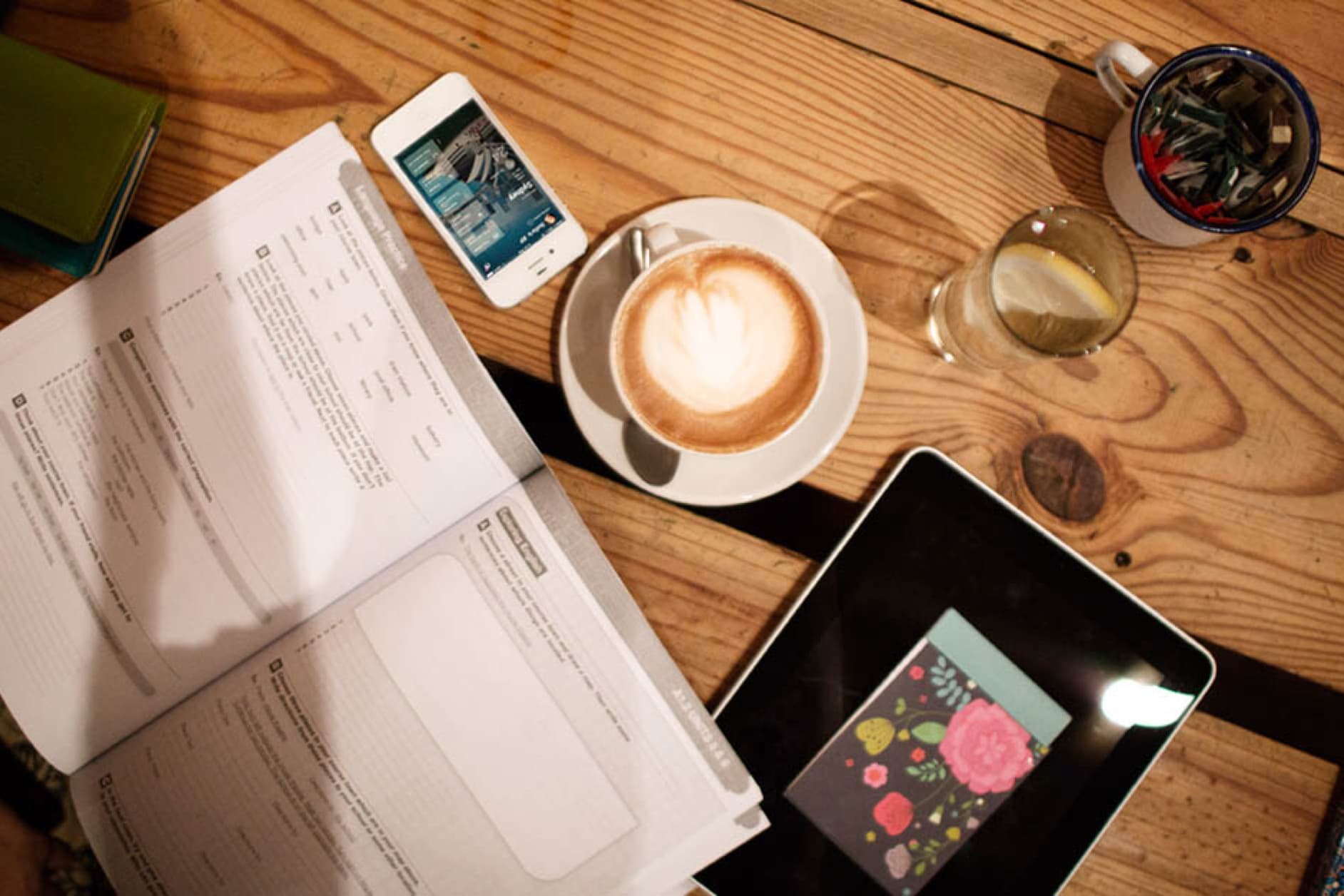 A cappuccino, smartphone, tablet, and some documents are laid out on a wooden table, ready for a productive session.