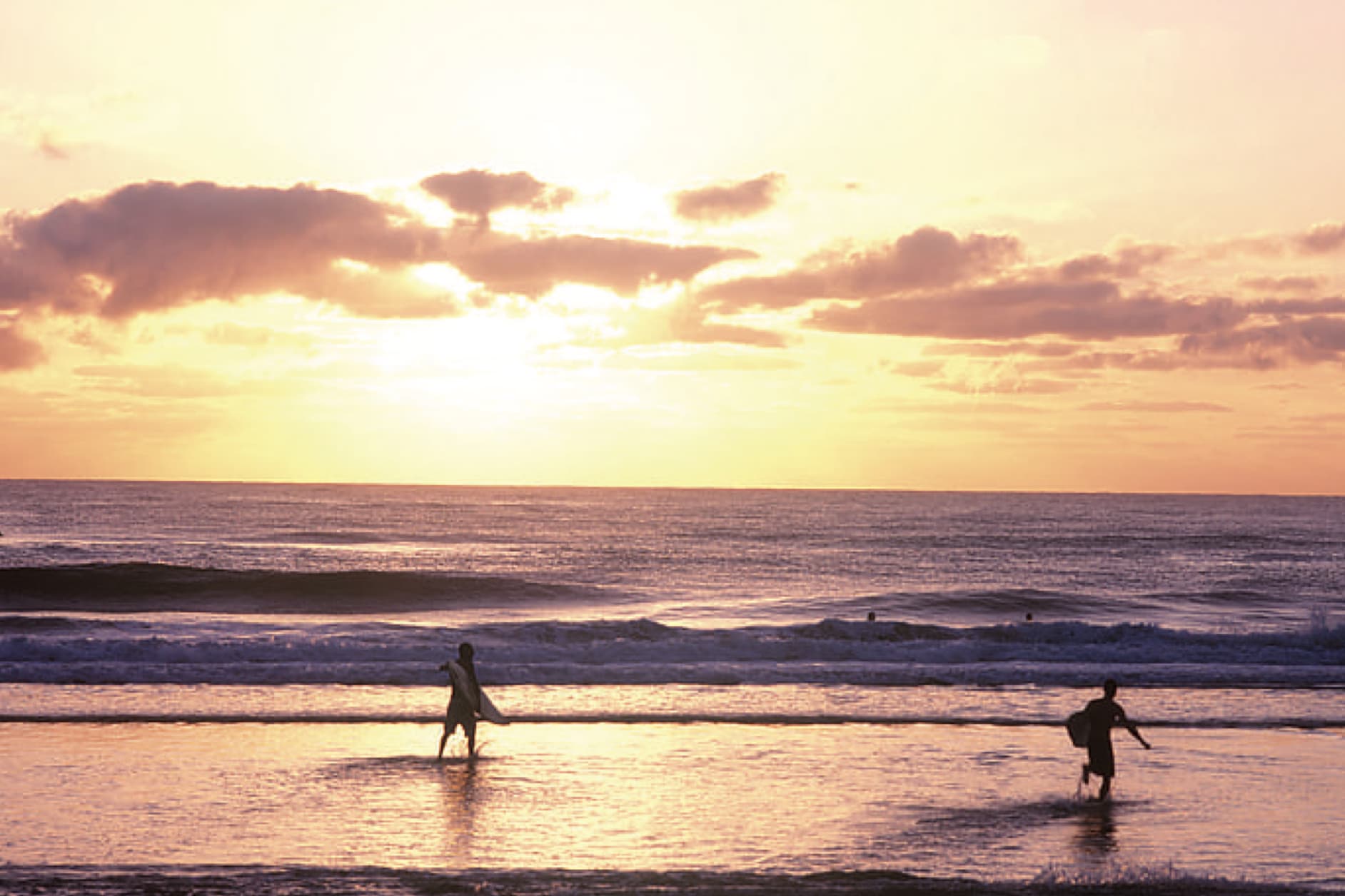 Two surfers walk into the ocean at sunset, with gentle waves and a warm, pastel-colored cloud cover painting the sky.
