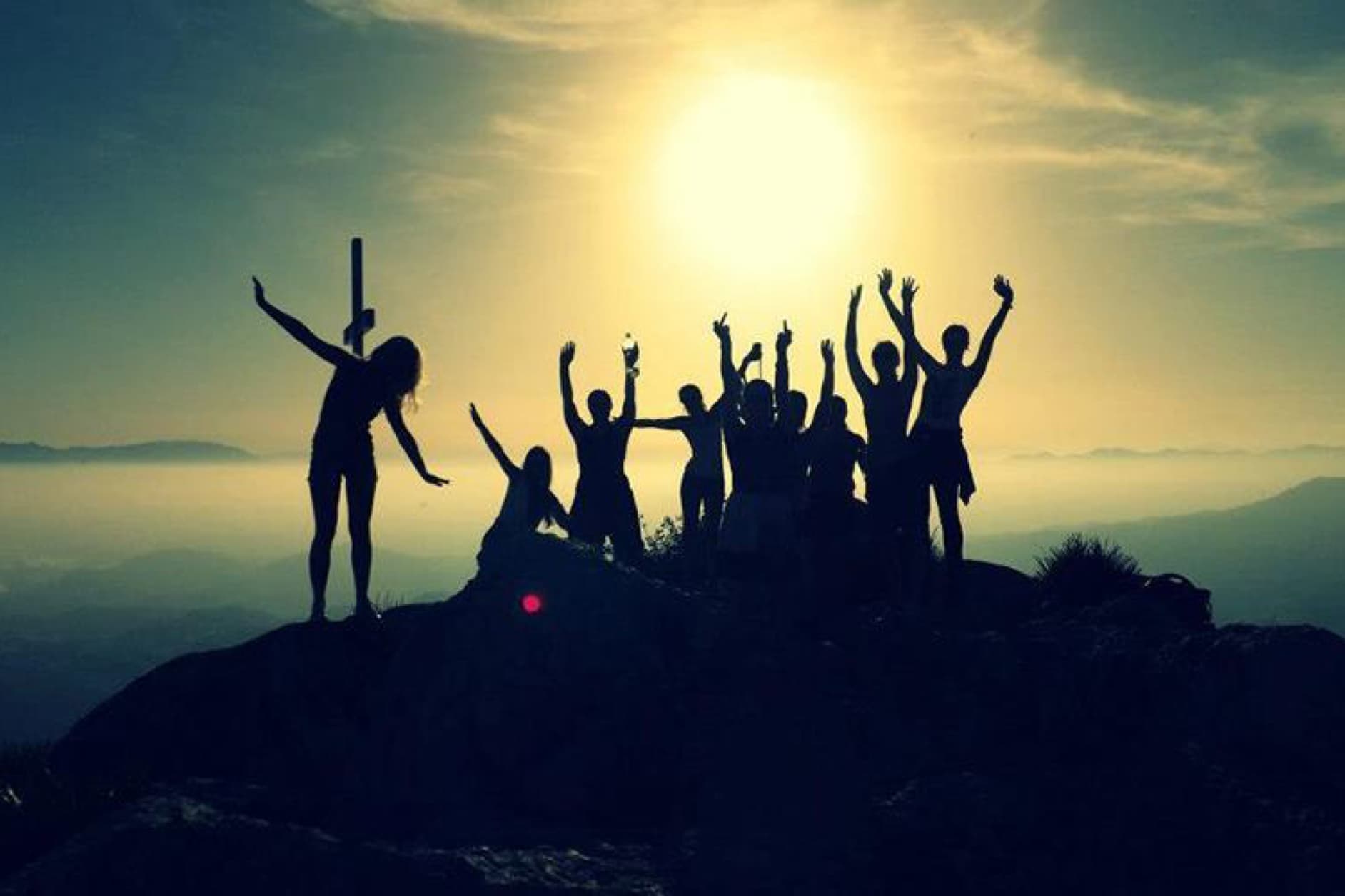 A group of friends celebrates on a mountaintop at sunset, with arms raised in the air, cheering for their achievement.