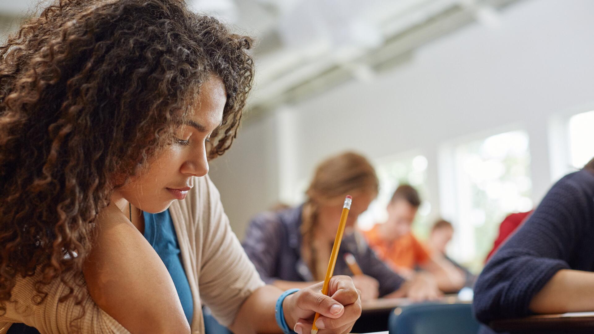 A young woman with curly hair is focused on writing during a test in a classroom setting, with other students blurred in the background.