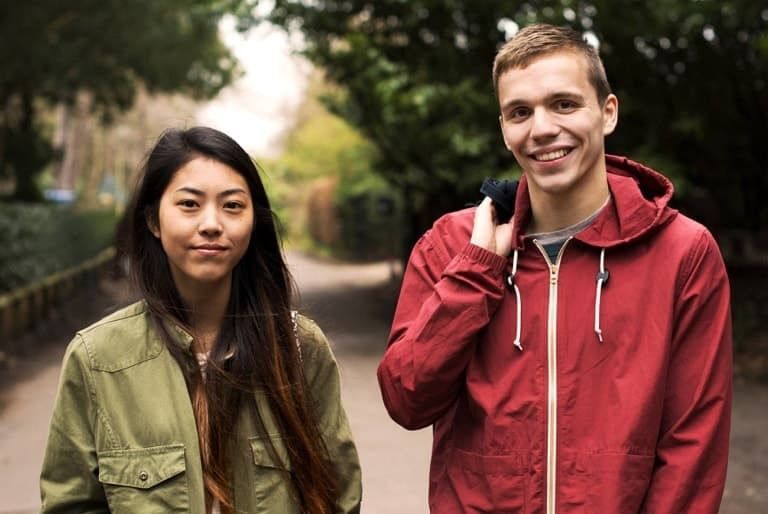 A young man and a young woman are walking side-by-side on a tree-lined path. The man is wearing a red hooded jacket, and the woman is wearing a green jacket.