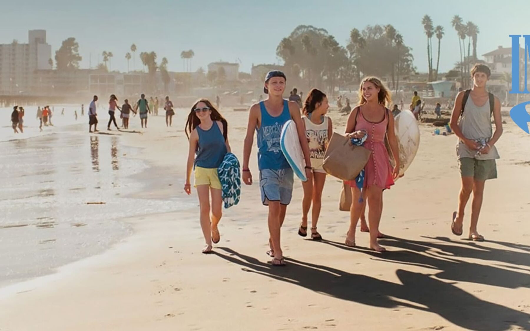 A group of young friends walk along a sunny beach, carrying surfboards and beach bags, with the ocean and distant buildings in the background.