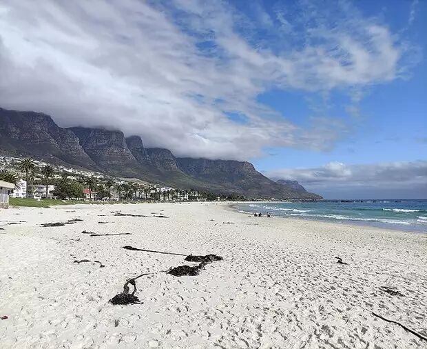 A scenic shot of a wide sandy beach with the turquoise ocean on the right and a range of mountains covered by clouds in the background.