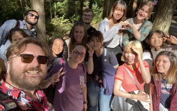 A diverse group of young adults are gathered outdoors in a sunny forest, smiling and posing for a selfie with peace signs.