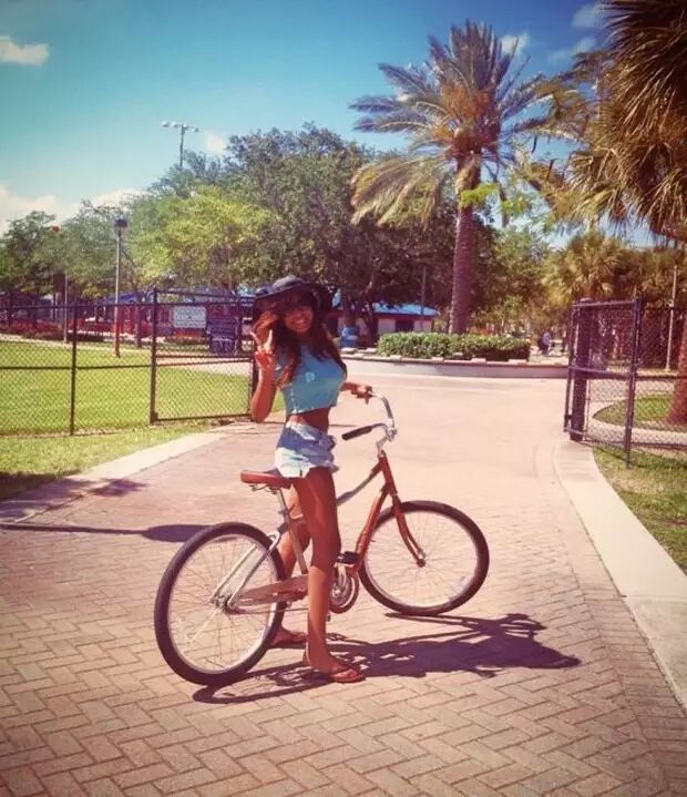 A young woman in a blue crop top and denim shorts poses with her bicycle on a sunny day, wearing a hat and giving a peace sign.
