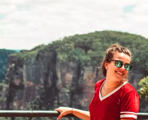 A woman wearing sunglasses and a red top smiles at the camera, posing in front of a stunning mountainous backdrop.