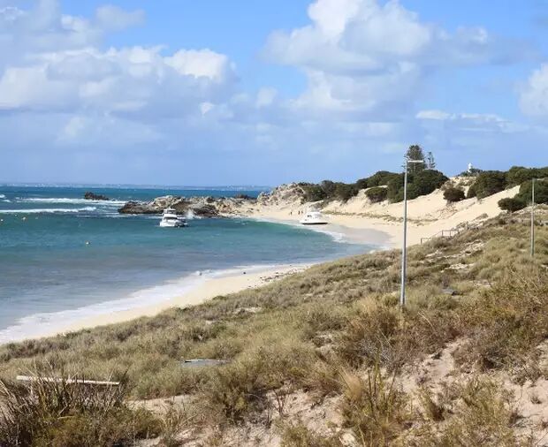 A scenic view of a sandy beach with turquoise waters, dotted with a few boats and lush green vegetation on the dunes under a partly cloudy sky.