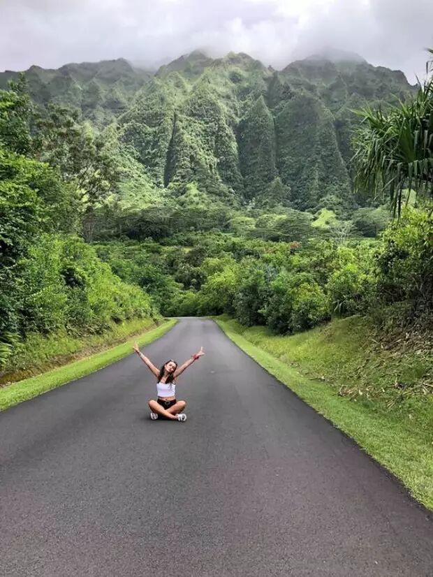 A woman sits on a road with her arms outstretched, facing a lush green mountainous landscape under a cloudy sky.