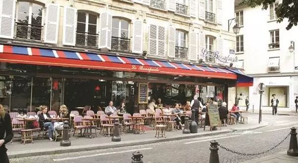 A bustling Parisian cafe with numerous patrons seated outdoors under red and blue awnings, enjoying the vibrant atmosphere.