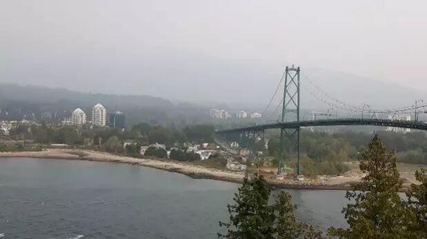 The image shows the Lions Gate Bridge in Vancouver, Canada, shrouded in a hazy, smoky atmosphere, likely due to wildfires. The water in the foreground reflects the overcast sky.