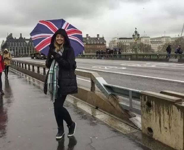 A woman holding a Union Jack umbrella stands on a wet street in London, with buildings visible in the background.