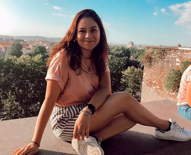 A young woman is sitting outdoors on a wall, smiling at the camera. She is wearing a pink top and striped shorts. A cityscape is visible in the background.